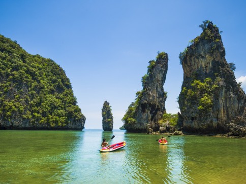 Two people in a kayak paddle through clear green water surrounded by tall, rocky limestone cliffs and lush greenery under a bright blue sky. Another kayak is visible in the distance.