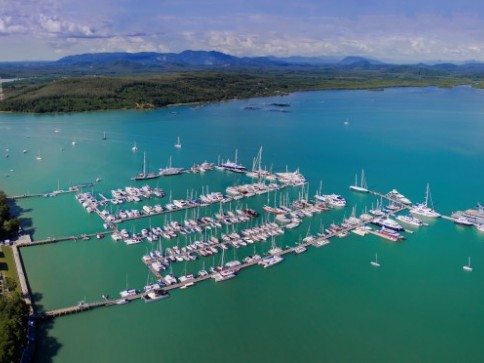 Aerial view of a marina with numerous boats and yachts moored in turquoise water, surrounded by green land and distant mountains under a partly cloudy sky.