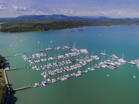 Aerial view of a marina with many yachts and sailboats moored in calm turquoise waters, surrounded by green shoreline and distant mountains under a partly cloudy sky.