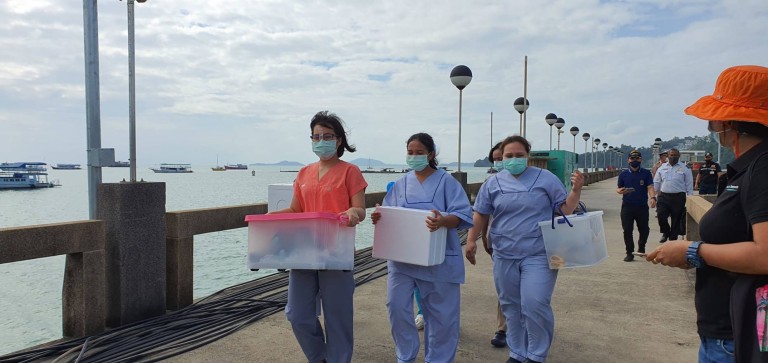 Three people wearing medical scrubs and face masks carry containers along a pier by the sea, accompanied by others. Boats are visible in the water and the sky is partly cloudy.