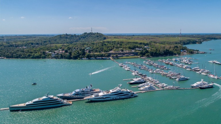 Aerial view of a marina with numerous yachts and boats moored along long piers, surrounded by green trees and calm blue-green water under a clear sky. A forested hill is visible in the background.