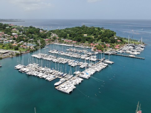 Aerial view of a marina with rows of moored sailboats and yachts in clear blue water, surrounded by greenery, coastal buildings, and a small peninsula extending into the sea.
