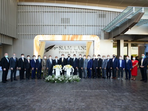 A large group of people in formal attire and face masks stand in front of a Siam Land Private Jet Terminal sign, posing for a group photo with a flower arrangement in the centre.
