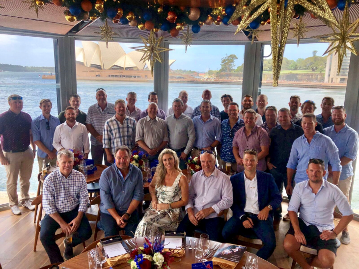 A large group of people, mostly men in casual and semi-formal attire, pose for a photo in a decorated indoor space overlooking water and the Sydney Opera House through tall windows.