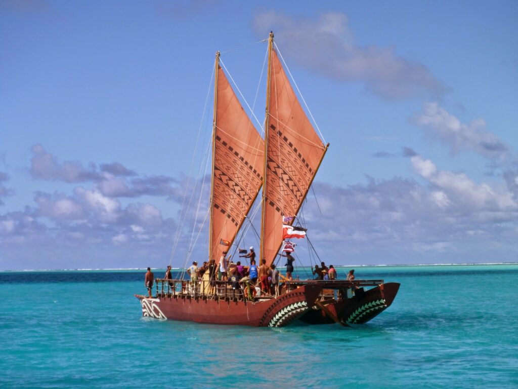 A traditional Polynesian double-hulled canoe with large reddish sails carries several people across turquoise ocean water under a blue sky with scattered clouds.
