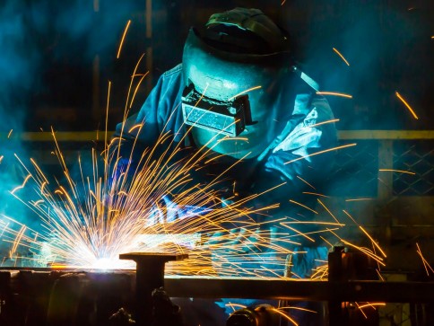 A person wearing a protective welding helmet and gloves is welding metal, with bright orange and yellow sparks flying in multiple directions against a dark industrial background.