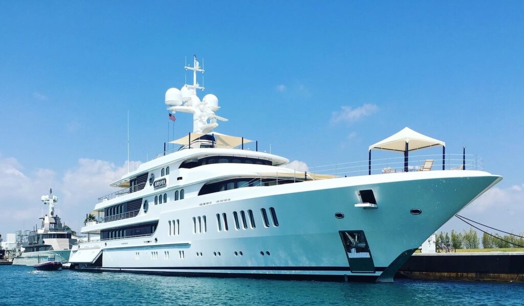 A large white luxury yacht is docked in a harbour on a sunny day, with clear blue sky and calm water. The yacht features multiple decks, shaded areas, and modern design elements.