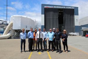Ten people stand in a row outside a large industrial building marked RIVERGATE.COM.AU, with boats and equipment visible in the background on a sunny day.