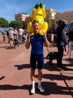 A smiling cyclist in a blue outfit stands outdoors, holding a large inflatable yellow kangaroo on his shoulders. Other people and bicycles are visible in the background on a sunny day.