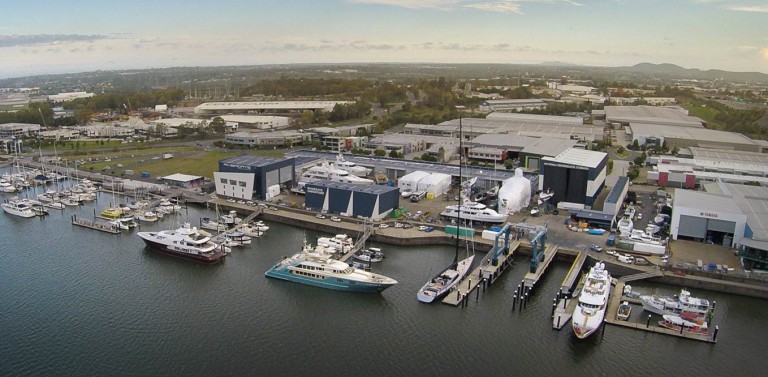 Aerial view of a marina with several yachts and boats moored along piers, surrounded by large industrial buildings and warehouses, with a cityscape in the background under a cloudy sky.