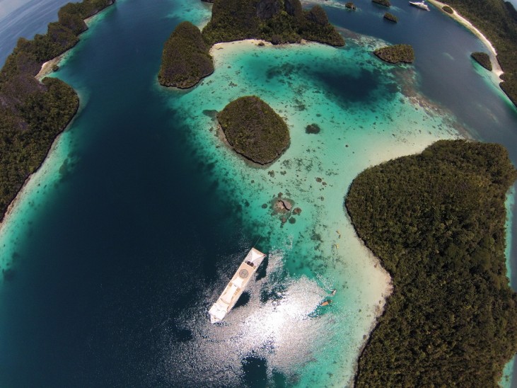 Aerial view of a white boat anchored in clear turquoise water surrounded by small, lush green islands and coral reefs. The sunlight reflects off the water near the centre of the image.
