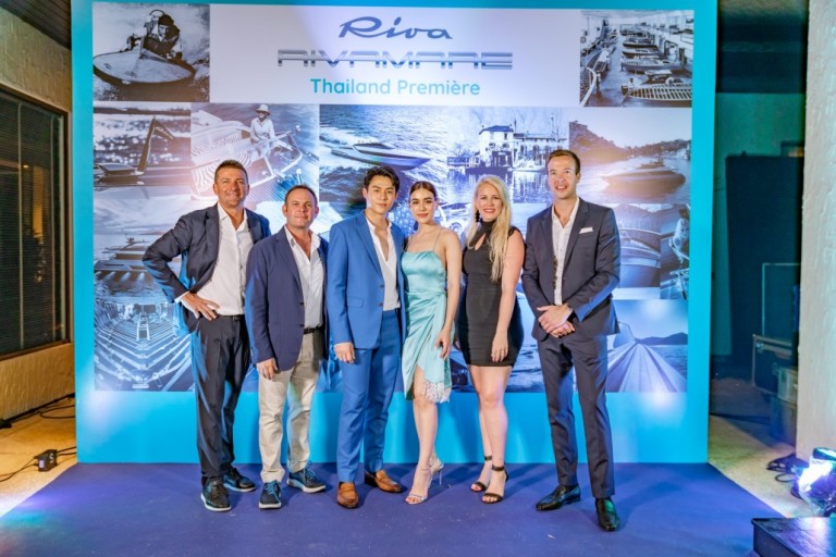 Six people dressed in formal attire pose together in front of a blue and white backdrop with Riva RIVAMARE Thailand Première and boating images at an event.