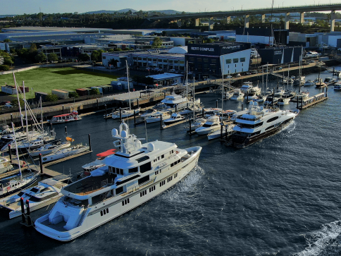 A large white yacht is leaving a marina filled with various yachts and boats. Industrial buildings and green fields are nearby, with a bridge and hills in the background. The scene is captured during daylight.