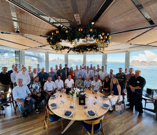 A large group of people pose for a photo around a round dining table set for a meal in a bright room with large windows, showing a waterfront view, the Sydney Opera House, and Sydney Harbour Bridge in the background.