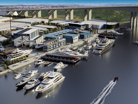 Aerial view of a modern marina with numerous yachts and boats moored along waterfront piers, adjacent to large industrial buildings, with a tall bridge and a city in the background.