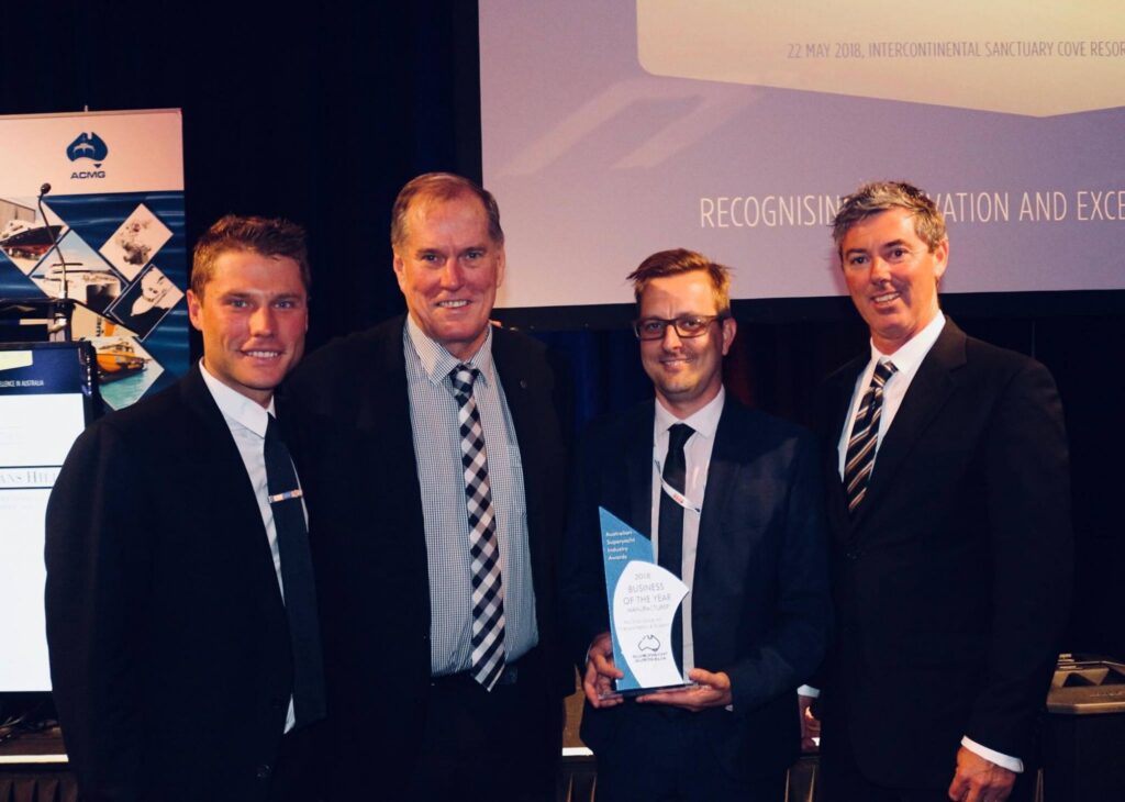 Four men in suits stand together indoors, smiling at the camera. One man in the centre holds a glass award. A projection screen and banners are visible in the background, suggesting a formal award ceremony or event.