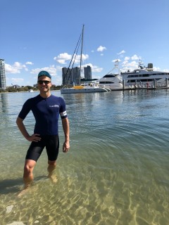 A man in a navy wetsuit stands in shallow water at a marina, with yachts and sailing boats moored behind him and tall buildings visible in the background under a blue sky.