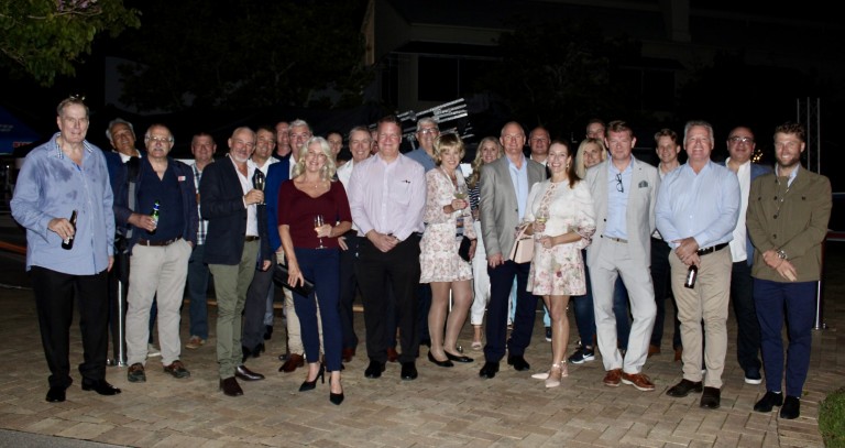 A group of around 25 adults, dressed in semi-formal attire, stand together outdoors at night, smiling for a group photo on a paved area, with trees and a building in the background.