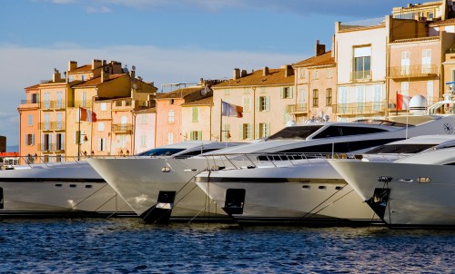 Luxury yachts moored in a marina with pastel-coloured buildings in the background, under a partly cloudy sky. The scene appears calm and picturesque, suggesting a popular waterfront destination.
