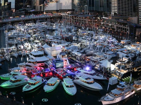 A marina packed with numerous yachts and boats, many illuminated with colourful lights, set against a backdrop of tall city buildings and waterfront structures at dusk.