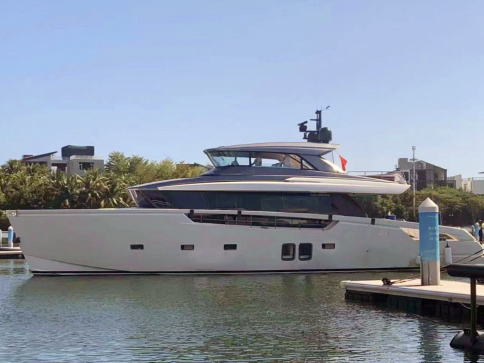 A modern white yacht is moored in a calm marina, with trees and contemporary buildings in the background under a clear blue sky.