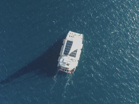 Aerial view of a white boat with solar panels on its roof sailing on deep blue water, casting a long shadow below.