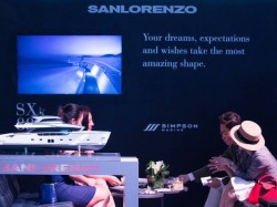 Four people sit at a table, talking, near a model yacht display. Behind them, a screen shows a yacht image. The wall reads: Your dreams, expectations and wishes take the most amazing shape.