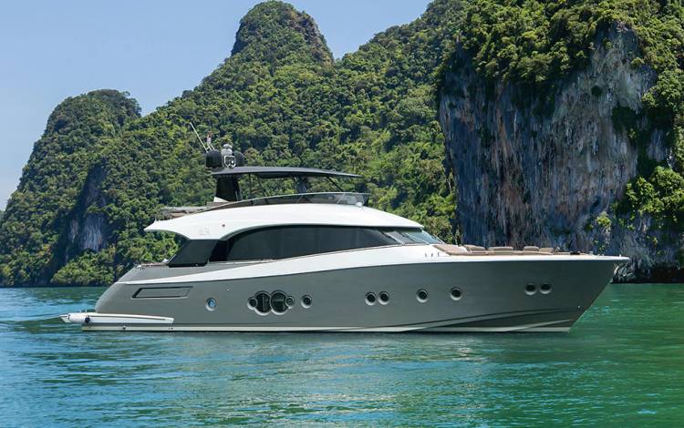 A sleek, modern yacht floats on clear turquoise water with lush, green cliffs and rocky hillsides in the background under a bright, sunny sky.