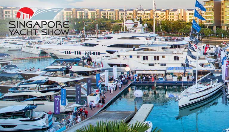 A vibrant marina scene at the Singapore Yacht Show with luxury yachts moored, visitors walking on the jetties, and the event logo displayed in the top left corner. Buildings and trees are visible in the background.