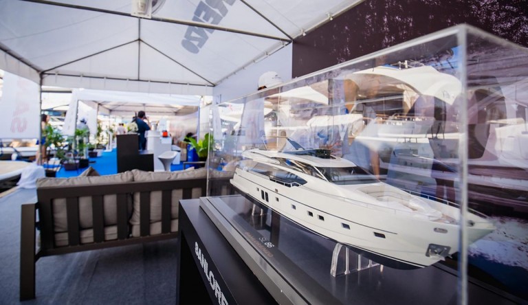 A detailed model yacht is displayed in a glass case at an indoor boat show, with people, seating, and banners visible in the background under a large marquee.