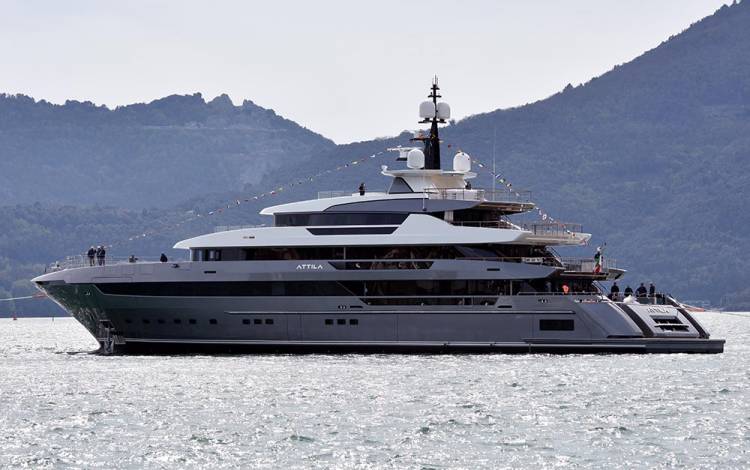 A large luxury yacht named Attila floats on calm water with mountains and greenery in the background under a clear sky.
