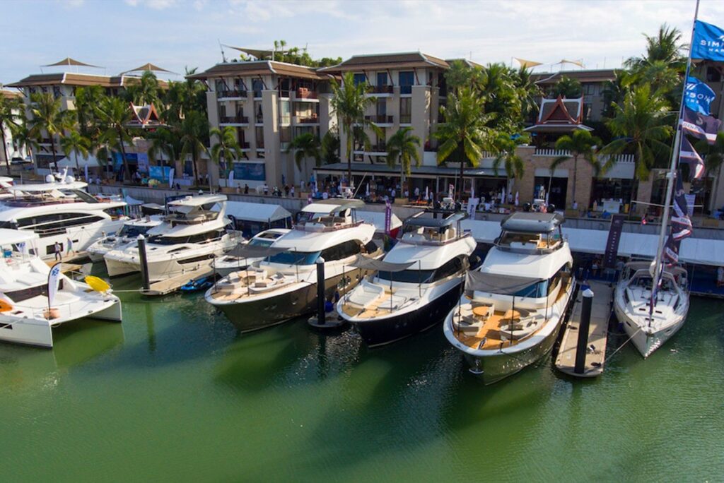 Several luxury yachts are moored side by side in a marina with green water, in front of a row of palm trees and upmarket resort-style buildings under a partly cloudy sky.