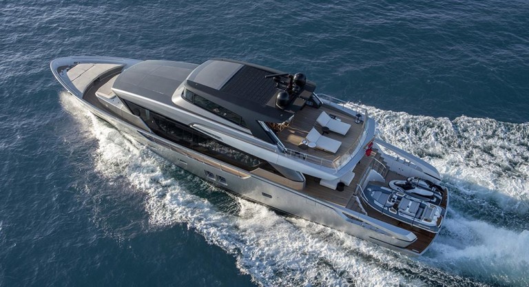 A modern luxury yacht cruises on blue water, leaving white waves behind. The yacht features spacious decks with lounge seating and a small boat stored at the stern.