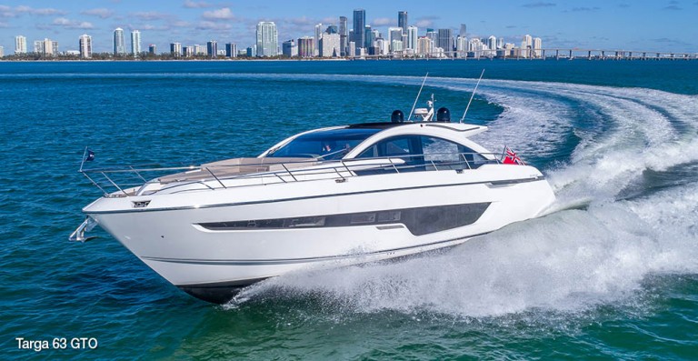 A sleek white yacht speeds across blue water, creating a trail of waves, with a city skyline and tall buildings visible in the background under a partly cloudy sky.