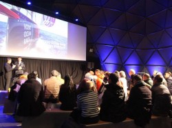 Audience seated in a dimly lit room watches two speakers on stage next to a large screen displaying an image and text, with blue lighting and geometric wall panels in the background.