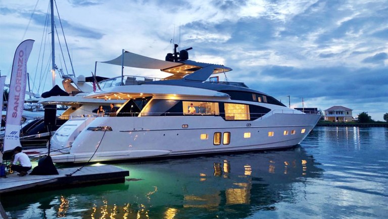 A luxury white yacht is moored at a marina during dusk, with lights illuminating the water below. The sky is cloudy, and nearby buildings and boats are visible in the background.