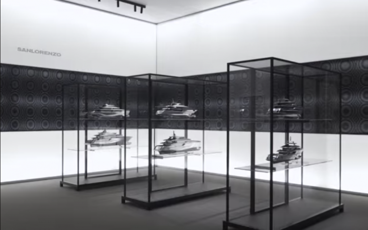 Glass display cases showcase detailed yacht models in a modern, minimalist showroom with white walls and geometric black patterns. The word SANLORENZO is visible on the left wall.