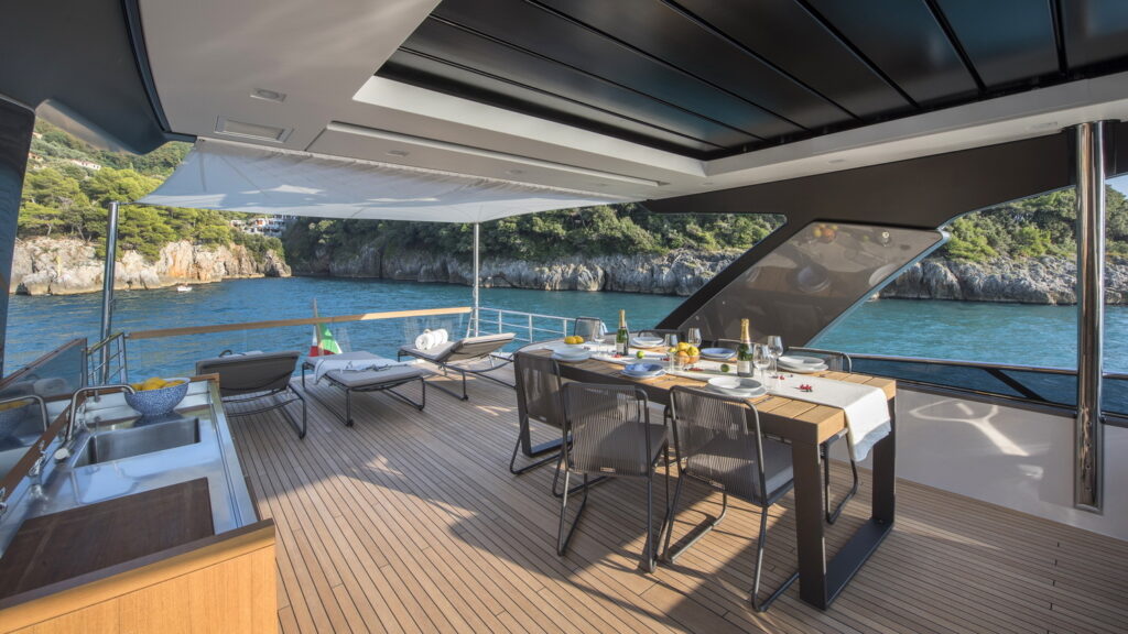 A luxury yacht deck with a dining table set for a meal, lounge chairs, a small kitchen area, and scenic views of blue water and rocky green cliffs in the background.
