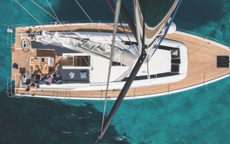 Aerial view of a sailboat with two people relaxing on the deck, sailing over clear turquoise water. The wooden deck and white sail of the boat are visible, surrounded by the sea.