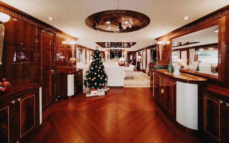 Luxurious yacht interior with rich wood flooring and panelling, a decorated Christmas tree with presents underneath, plush white seating, and large windows letting in natural light.