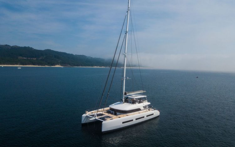 A white catamaran yacht floats on calm blue water near a distant shoreline with green hills under a partly cloudy sky.