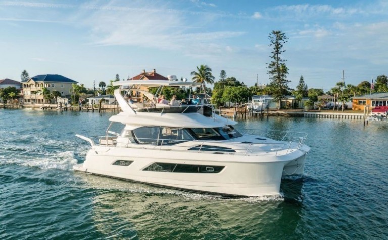 A white yacht cruises on calm water near a dock lined with houses and palm trees under a blue sky with scattered clouds. Several people relax on the yacht's upper deck.
