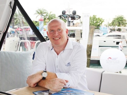A smiling man in a white shirt sits at a table on a boat with brochures in front of him. Yachts, palm trees, and show banners are visible in the background, suggesting a boat show or marina event.