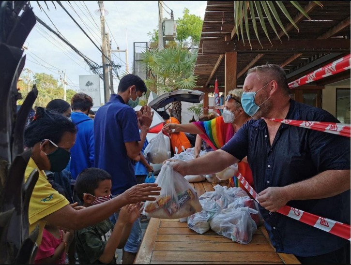 A group of people wearing masks distribute bags of food to others standing in a queue outdoors. The distribution is organised along a wooden table, separated by red tape, and everyone appears to be cooperating.