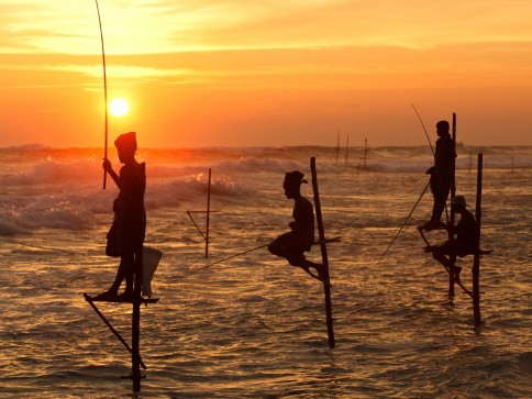 Silhouetted fishermen sit on wooden stilts above the ocean waves, casting their lines at sunset, with a vibrant orange sky and sun on the horizon.