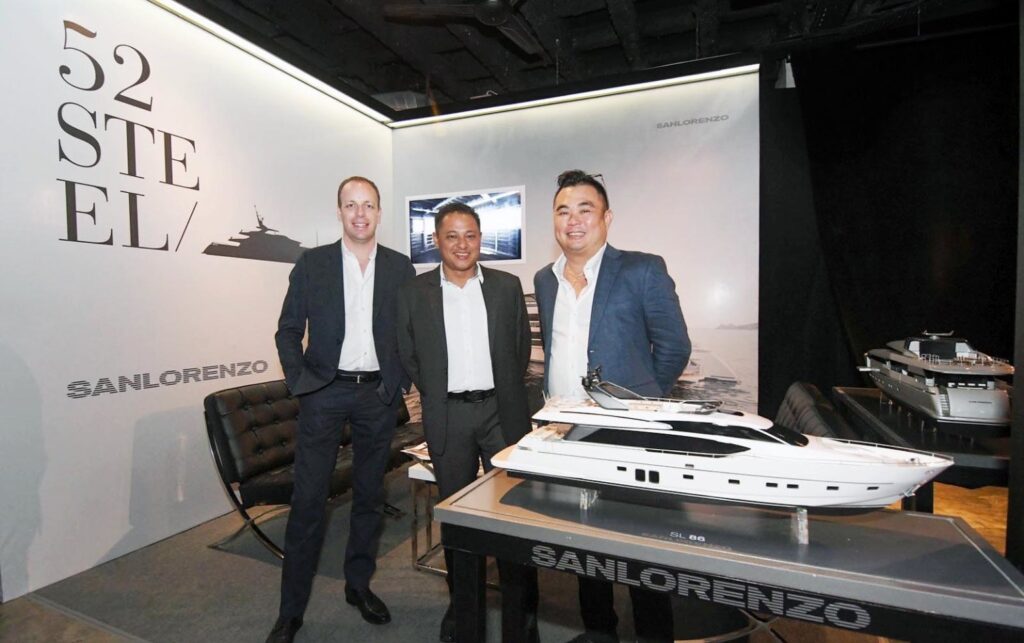 Three men in suits stand and smile beside a detailed model yacht at a Sanlorenzo stand, with 52 Steel displayed on the wall behind them and additional yacht models visible.