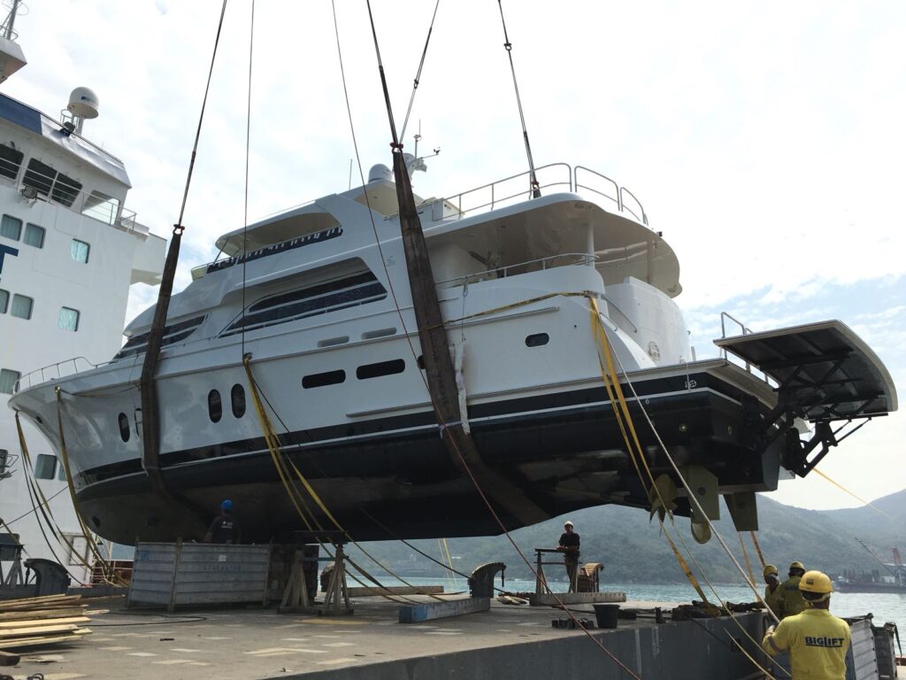 A large yacht is being lifted by cranes with straps at a dock. Workers in yellow safety helmets and shirts stand nearby, guiding the process. Mountains and water are visible in the background.