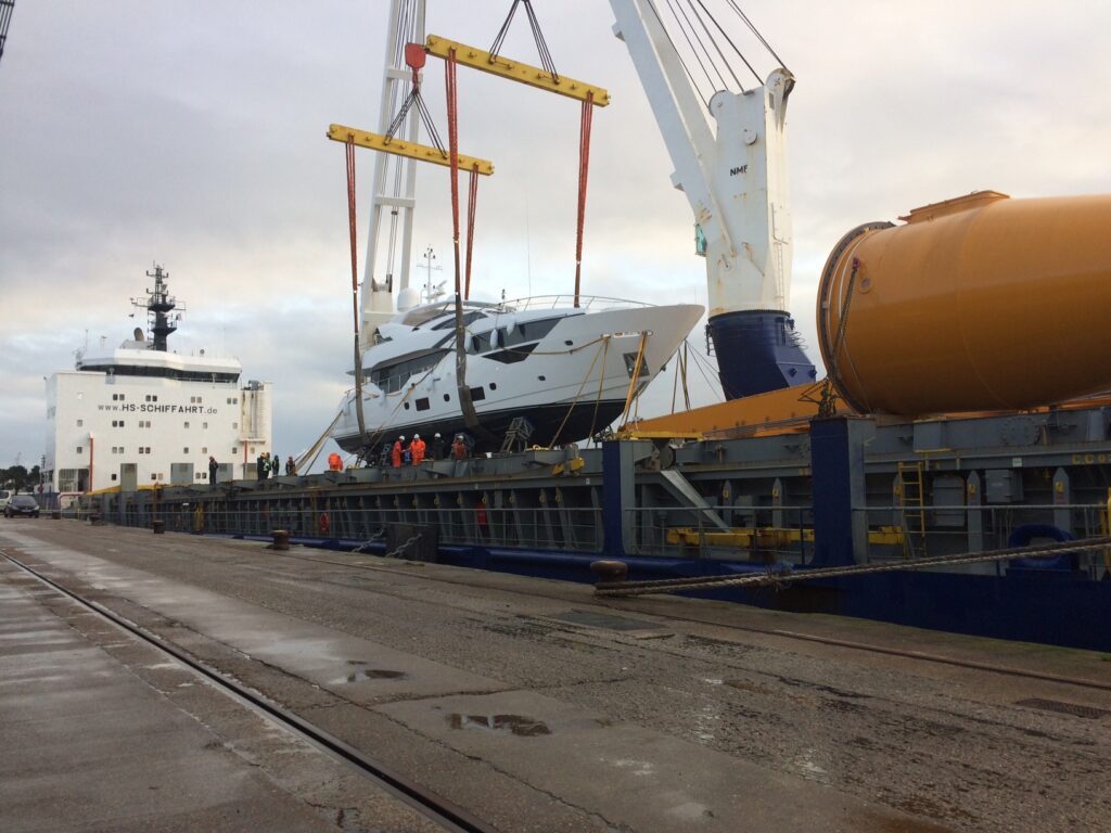 A large yacht is being lifted by cranes from a cargo ship at a quay. Several workers in safety vests are gathered nearby, and the area is wet with puddles on the ground.