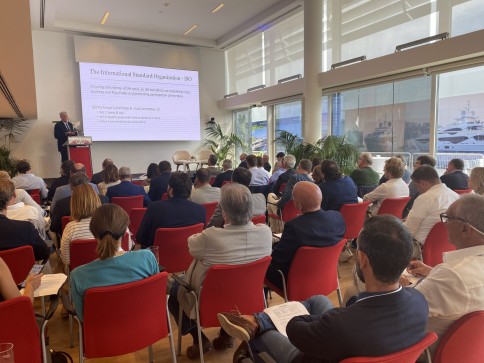 A man gives a presentation to an audience seated in red chairs in a modern, sunlit conference room. A large screen displays information about the International Organisation for Standardisation (ISO).