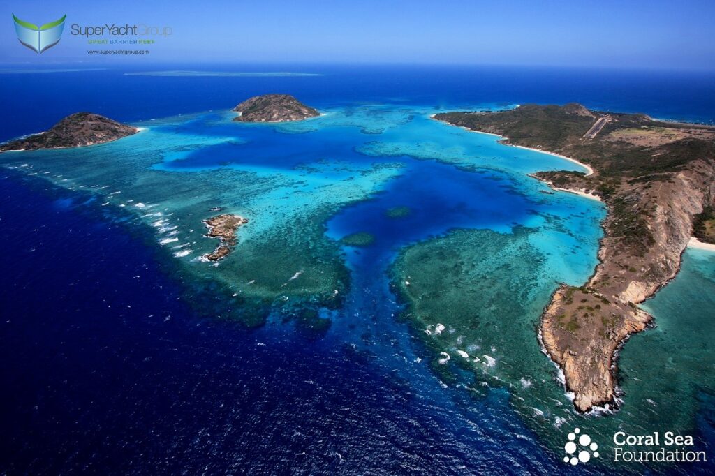 Aerial view of a tropical island surrounded by clear blue sea, coral reefs, and white sandy beaches, with lush green hills and a winding coastline. Logos for SuperYacht Group and Coral Sea Foundation appear in the corners.